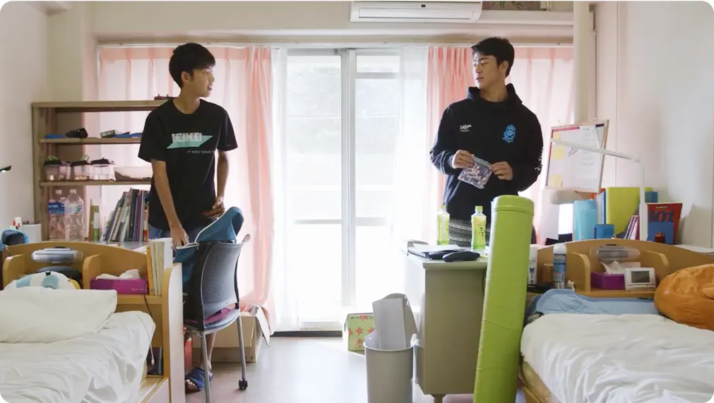 기숙사 방에서 학생 두 명이 이층 침대와 책상 사이에 서 있는 모습