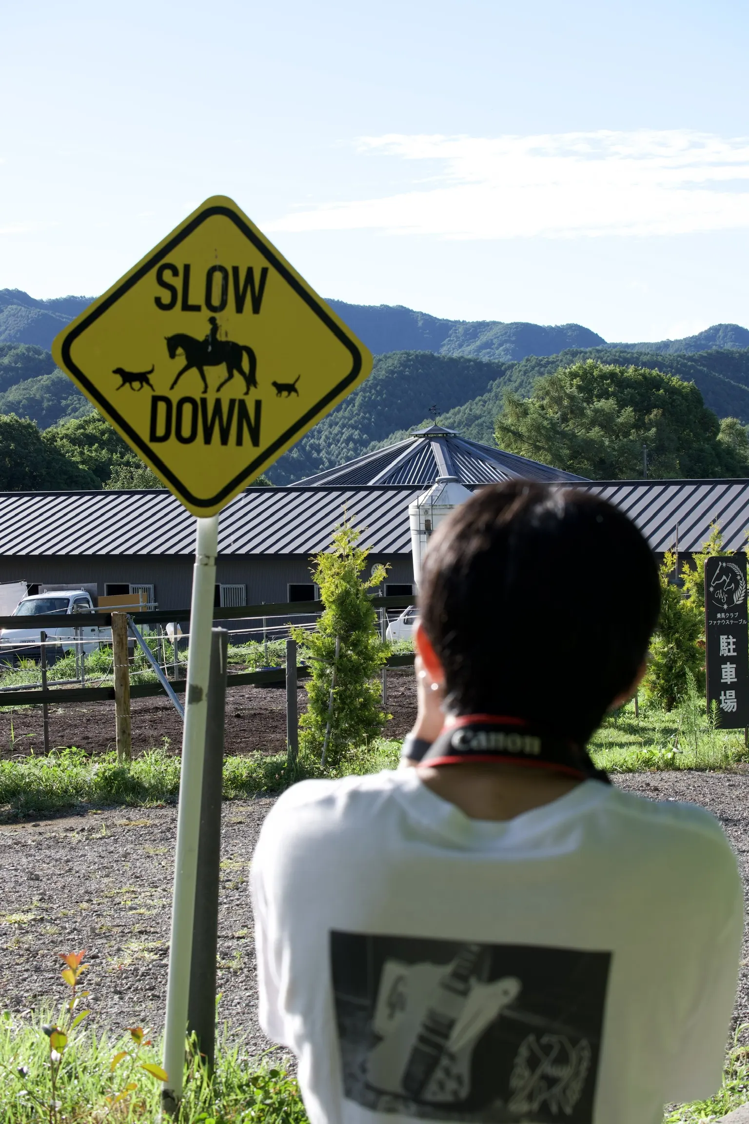 SLOW DOWN 노란 표지판 너머 산자락의 승마 클럽을 카메라로 찍는 사진부 학생의 뒷모습