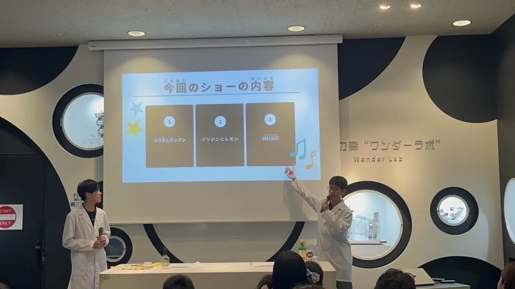 원더 랩 무대에서 흰 가운을 입고 마이크를 든 채 스크린의 실험 쇼 순서를 설명하는 화학반 학생들
