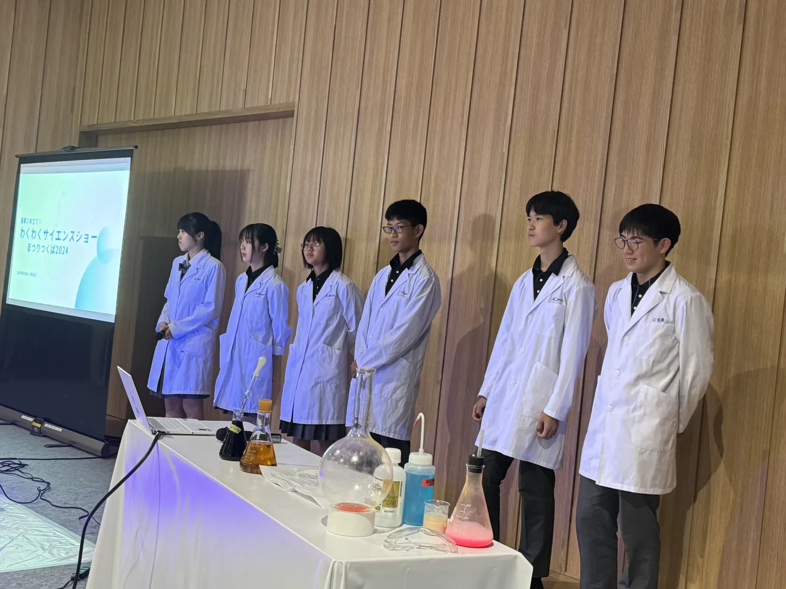 마쓰리 쓰쿠바 2024 사이언스 쇼에서 플라스크와 시약병을 늘어놓은 탁자 뒤에 나란히 선 흰 가운 차림의 화학반 학생들