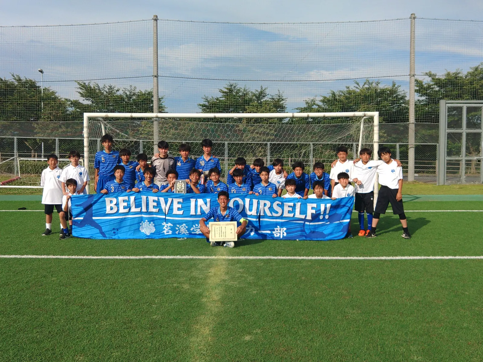 골대 앞에서 BELIEVE IN YOURSELF 현수막과 상장, 트로피를 들고 찍은 중학 축구부 단체 사진