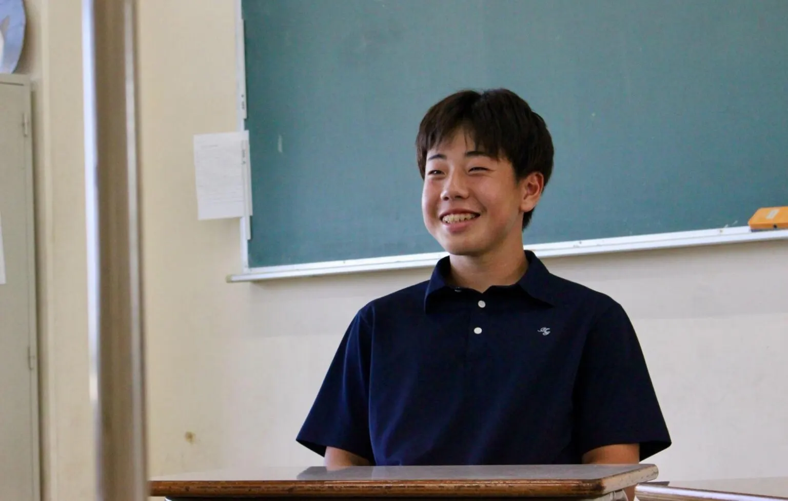 교실 칠판 앞 책상에 앉아 활짝 웃는 남색 폴로셔츠 차림의 R 학생