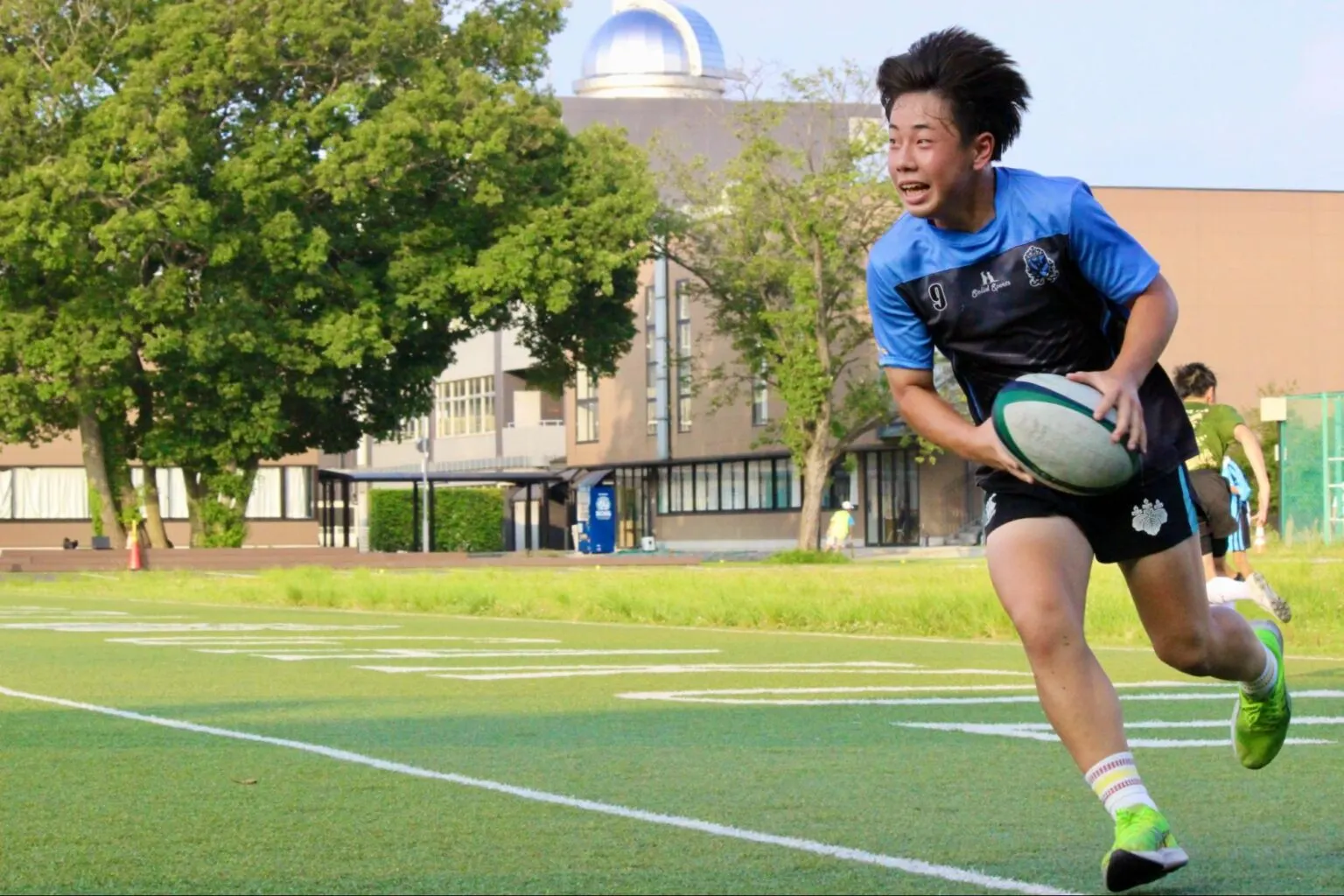돔 지붕 관측소가 있는 교사 앞 인조 잔디 운동장에서 럭비공을 들고 달리는 등번호 9번 R 학생