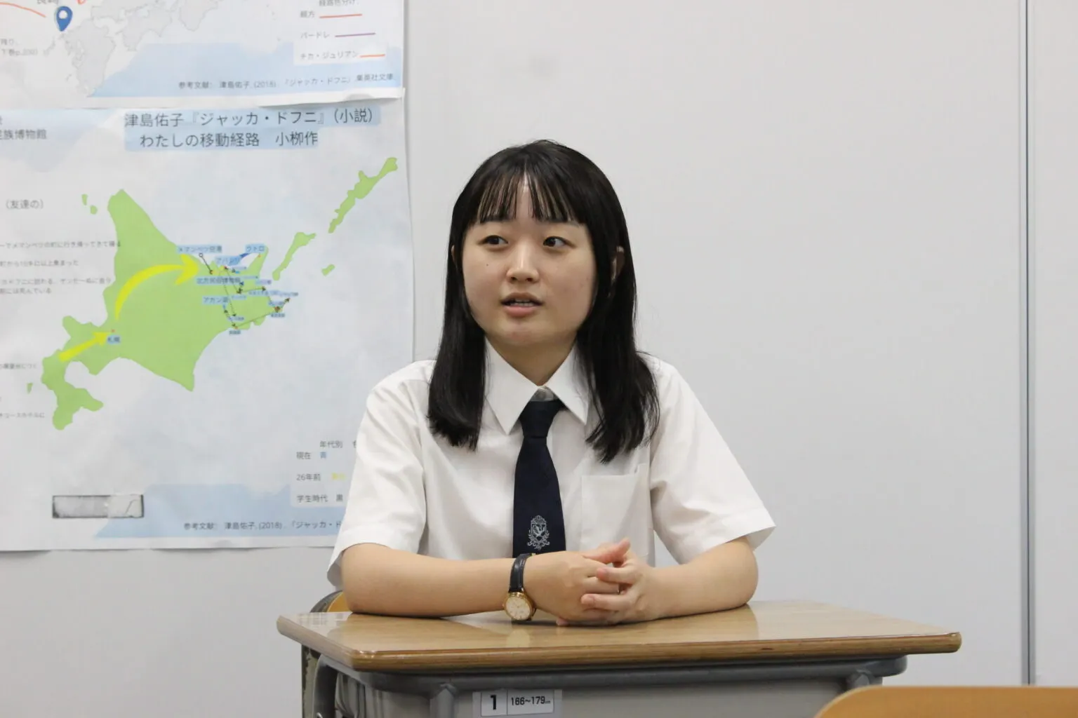 홋카이도 지도가 그려진 게시물 옆 책상에 두 손을 모으고 앉아 이야기하는 I 학생