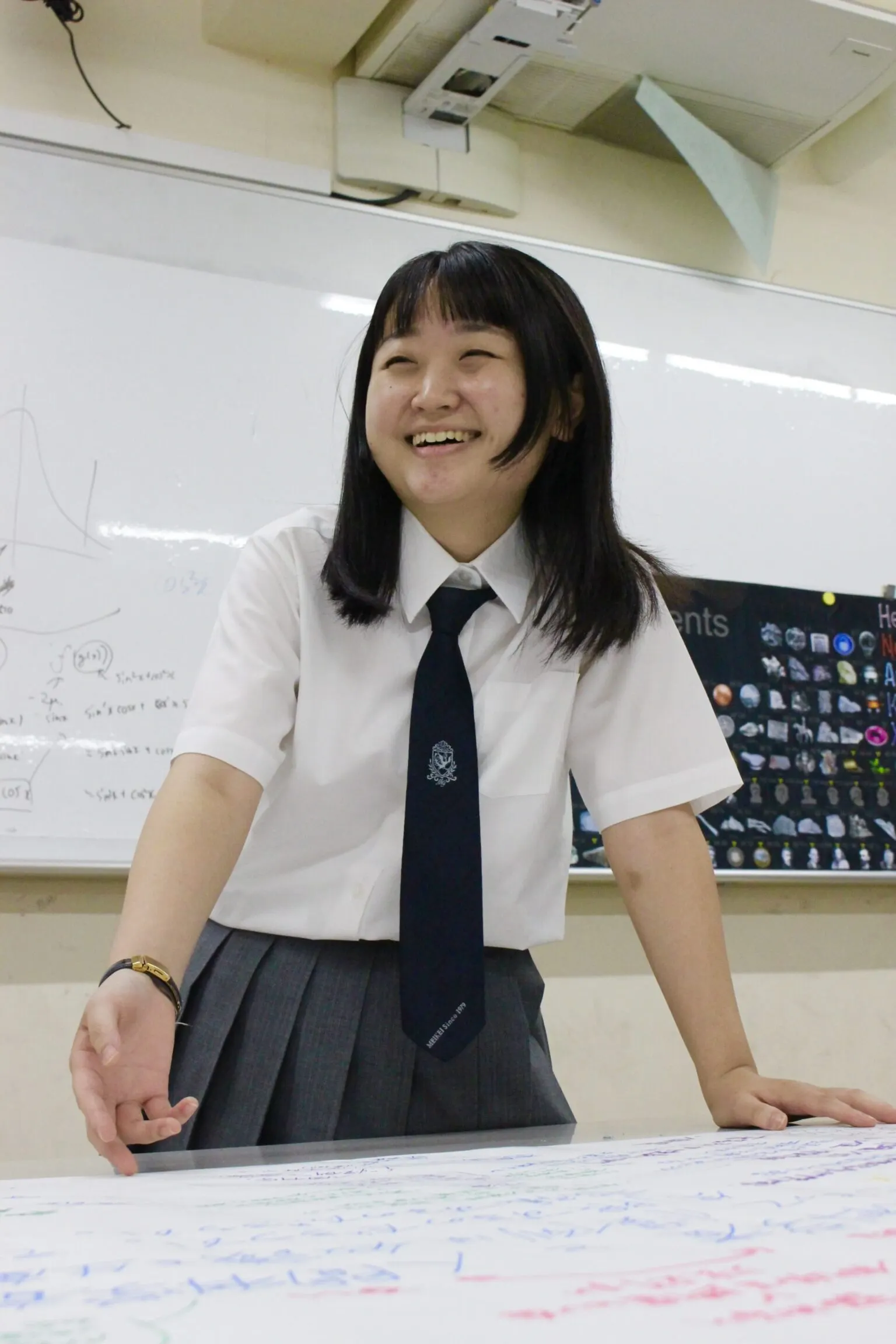 수식이 적힌 화이트보드 앞에서 손글씨가 가득한 큰 종이가 놓인 책상에 손을 짚고 활짝 웃는 I 학생