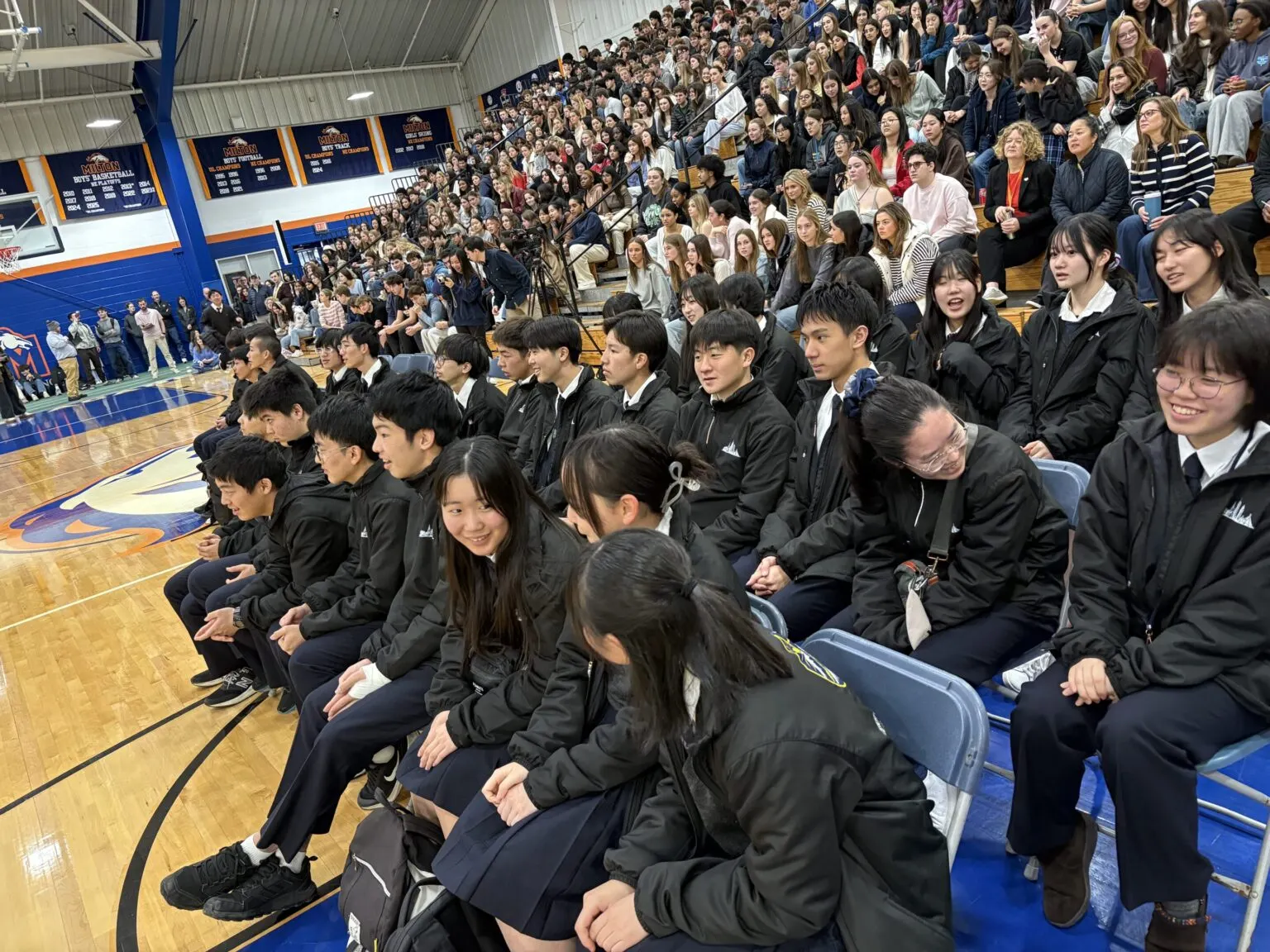 밀턴 아카데미 체육관에서 현지 학생들로 가득 찬 관중석 앞줄 의자에 나란히 앉은 메이케이가쿠엔 고등학교 학생들