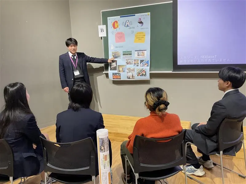 칠판에 붙인 CAS 포스터를 가리키며 의자에 앉은 학생들에게 설명하는 교복 차림 남학생