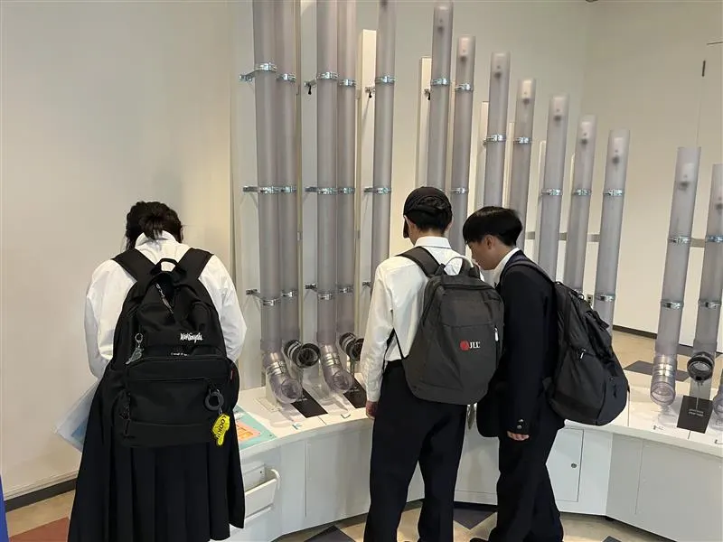 벽면에 세운 여러 개의 회색 관 모양 과학 전시물을 살펴보는 학생 세 명의 뒷모습
