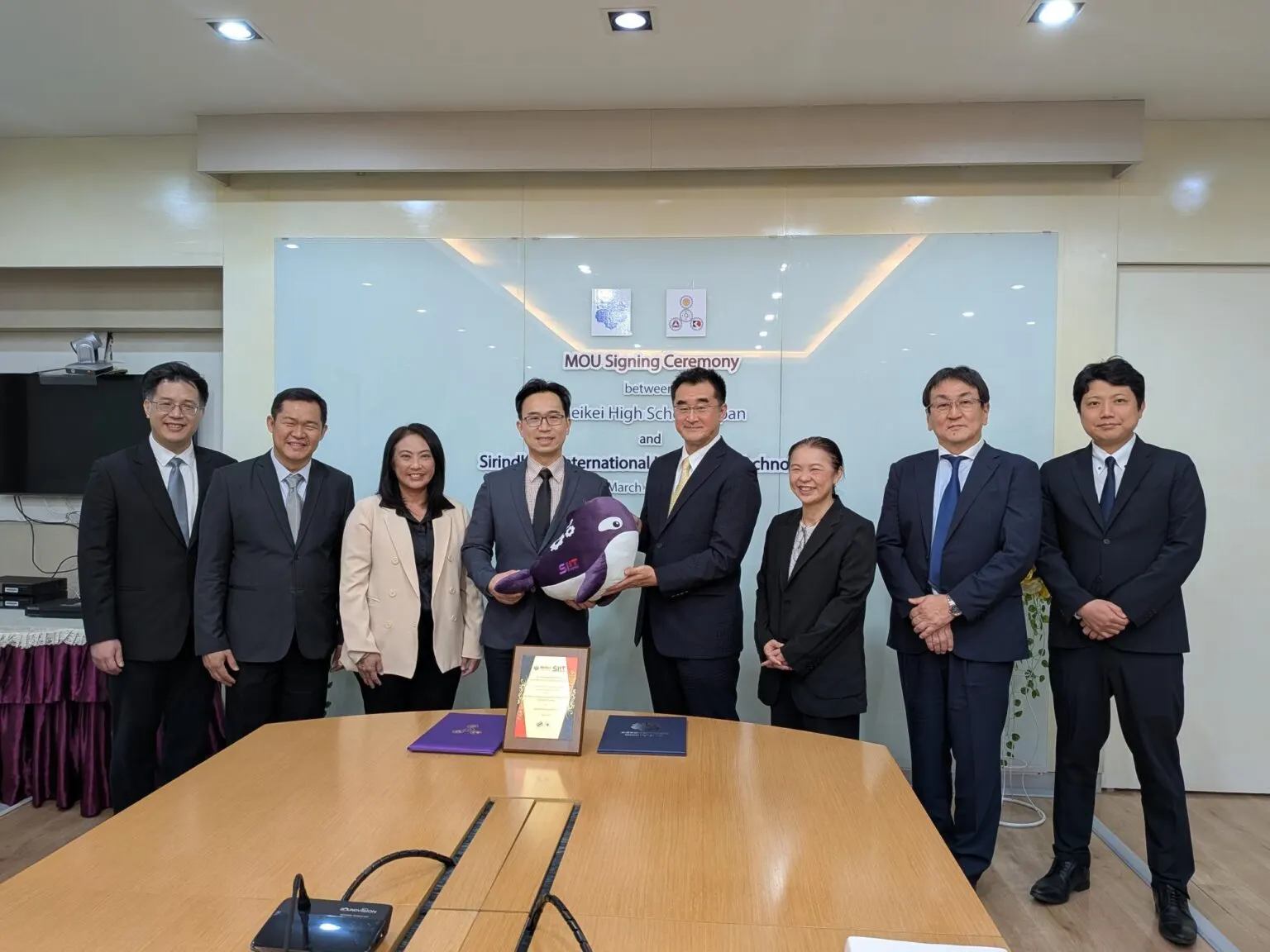 MOU 서명식장에서 양교 관계자 8명이 나란히 서고, 가운데 두 대표가 SIIT 로고가 달린 보라색 범고래 인형을 함께 들고 있는 기념사진