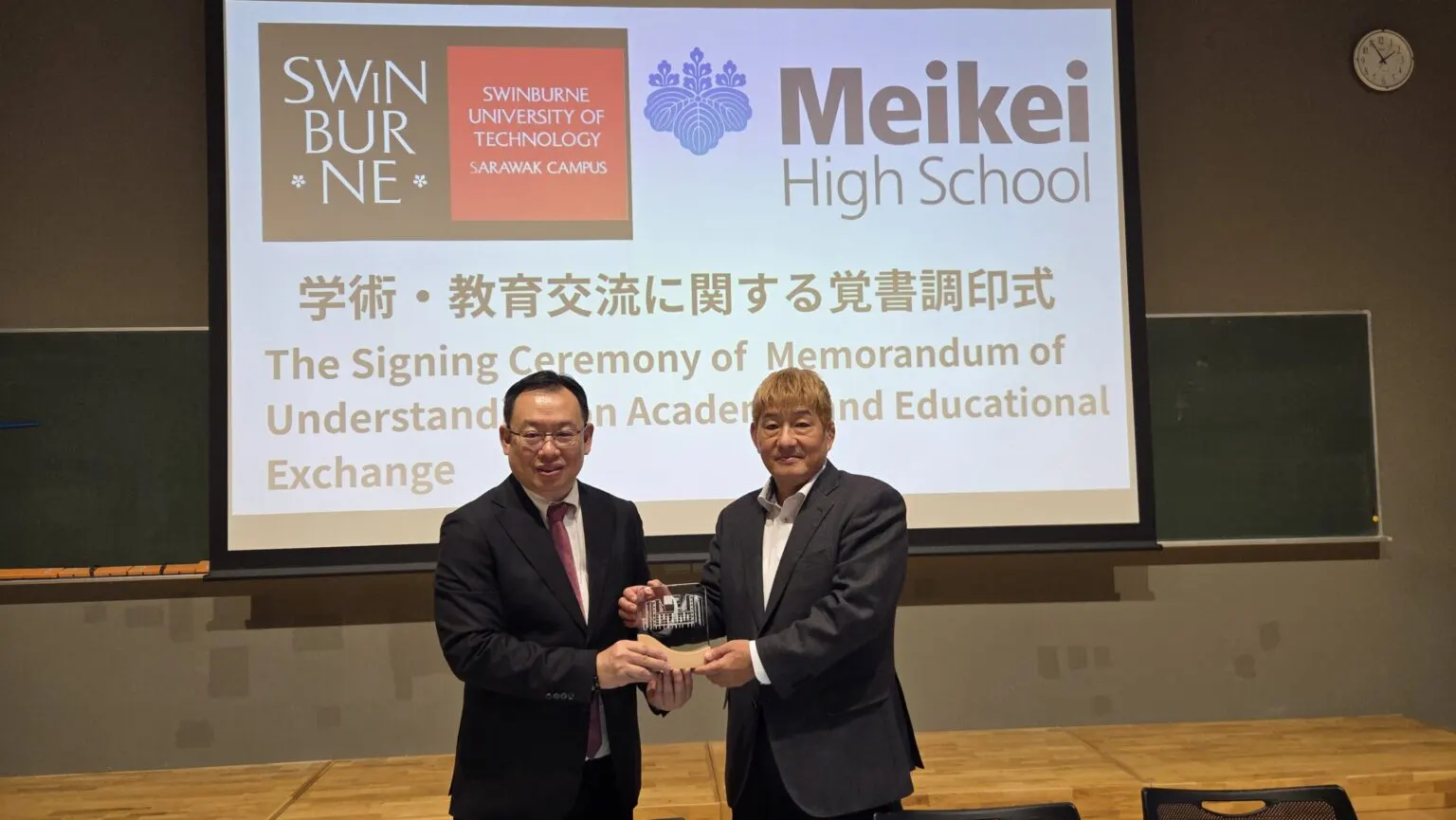 조인식 화면 앞에서 기념패를 함께 들고 선 Swinburne University of Technology Sarawak 대표와 메이케이가쿠엔 고등학교 대표