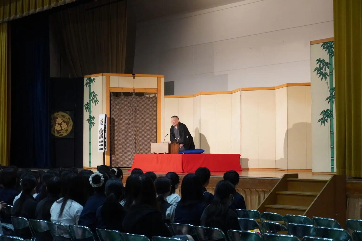 대나무 그림 병풍을 두른 체육관 무대의 붉은 고좌에 앉아 라쿠고를 하는 검은 하오리 차림의 라쿠고가와 객석의 학생들