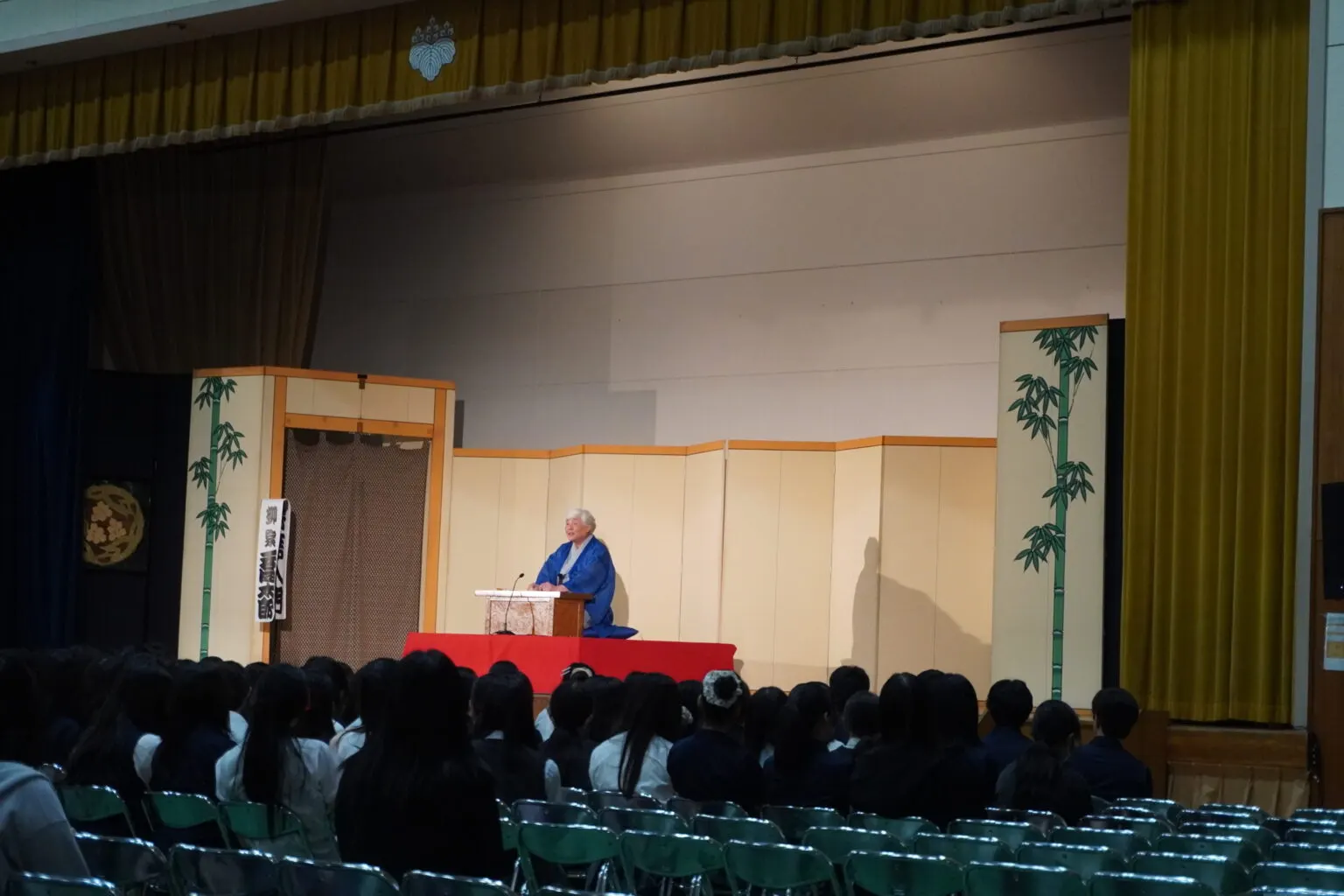 파란 기모노 차림으로 무대 위 고좌에 앉아 라쿠고를 하는 라쿠고가와 이를 바라보는 학생들