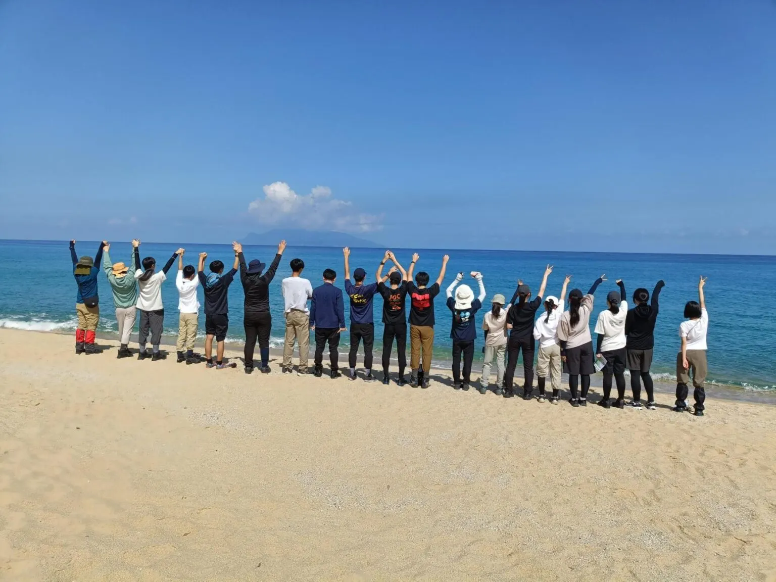파란 바다를 바라보며 모래사장에 한 줄로 서서 두 팔을 높이 든 참가 학생과 인솔자들의 뒷모습