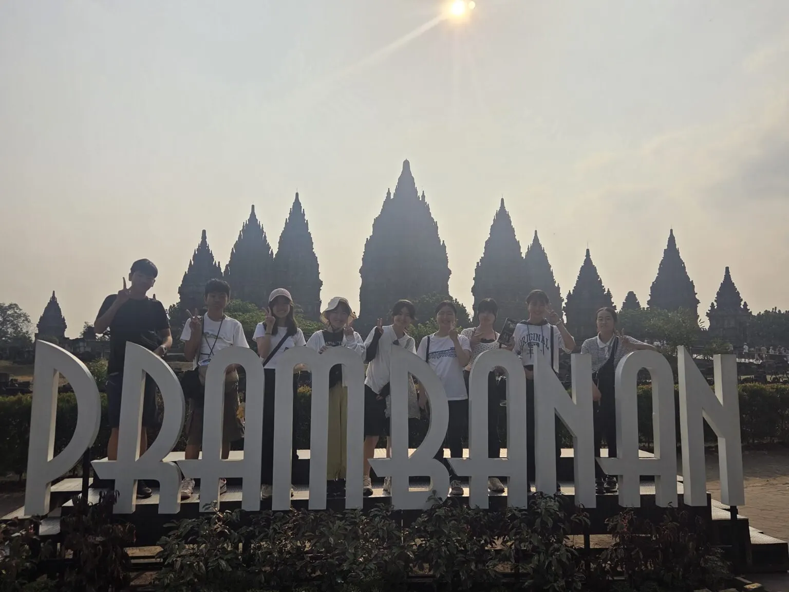 프람바난 사원의 탑들을 배경으로 PRAMBANAN 글자 조형물 뒤에 서서 손가락 브이를 한 연수 참가 학생 9명