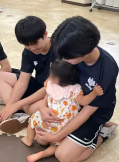 바닥 매트에 앉아 꽃무늬 원피스를 입은 아기를 무릎에 앉히고 들여다보는 남학생 두 명