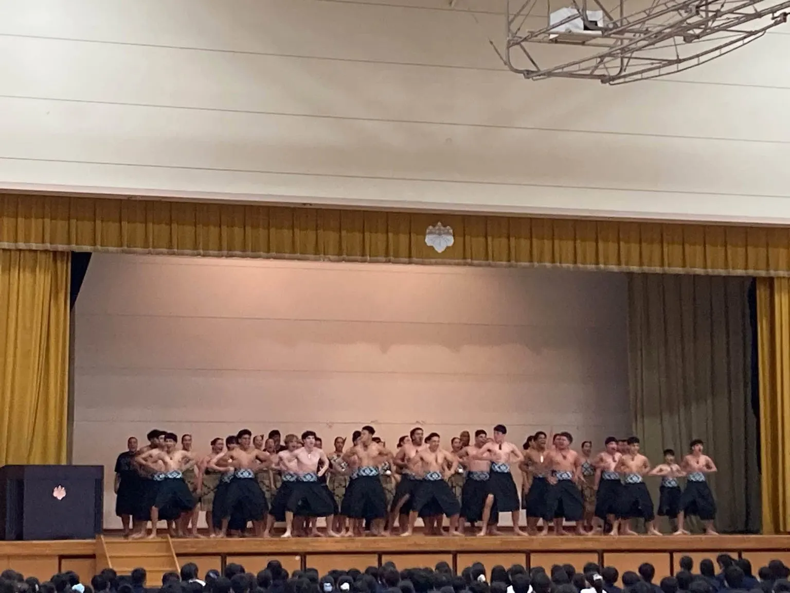 체육관 무대에서 웃옷을 벗고 검은 치마형 의상을 입은 남학생들이 앞줄에서 힘차게 하카를 추는 모습