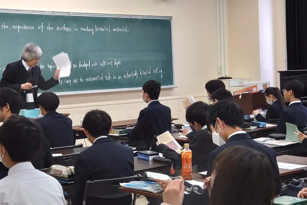 강의실에서 교원이 영어 판서 앞에서 설명하고 학생들이 자료를 보는 모습